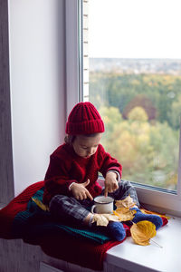 Boy a child in a red sweater and a knitted hat sits at the window on the windowsill