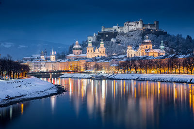 Illuminated buildings by lake against sky at dusk