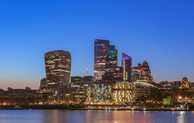 Illuminated buildings by river against clear sky