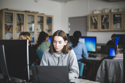 Confident female student using laptop at desk against teacher and friends sitting in computer lab