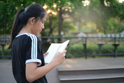 Side view of girl reading book while standing against trees in park