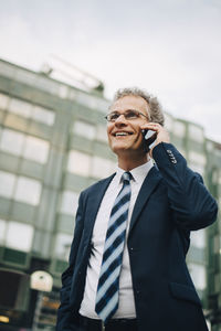 Low angle view of smiling businessman talking through smart phone while standing in city