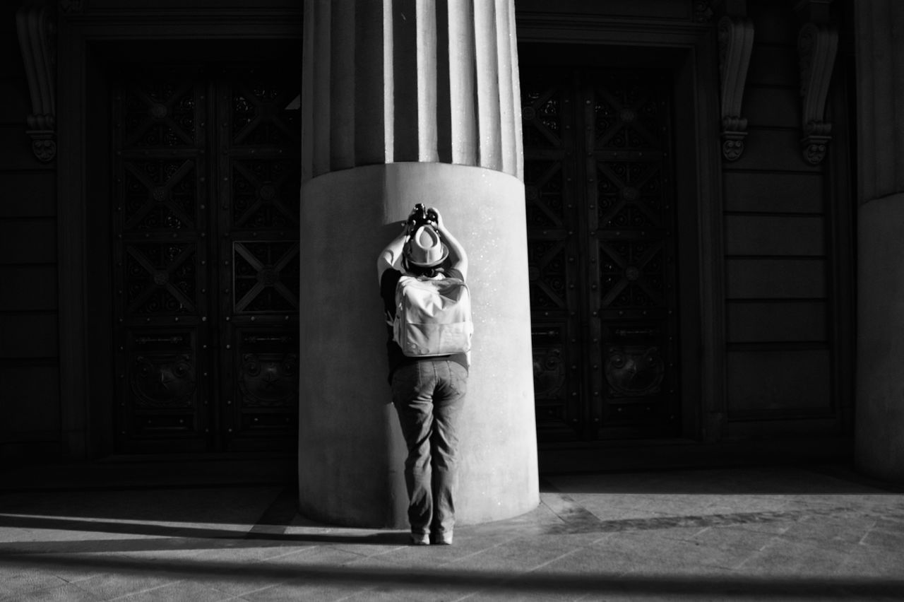 black, white, black and white, darkness, monochrome, architecture, monochrome photography, one person, full length, built structure, entrance, building exterior, door, light, adult, standing, clothing, building, person, day, lifestyles, women, architectural column, sunlight, outdoors, men, shadow