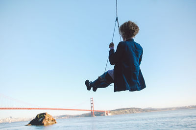 Low angle view of woman swinging over sea against sky with golden gate bridge in background