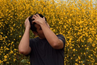 Young man with hands in hair standing against yellow flowering plants at farm