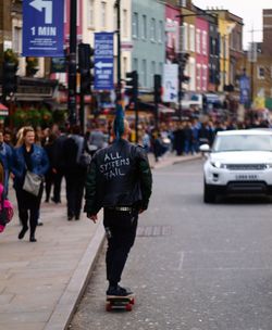 Rear view of hipster skateboarding by sidewalk on street in city