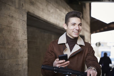 Smiling teenage boy with electric push scooter looking away while holding mobile phone below bridge