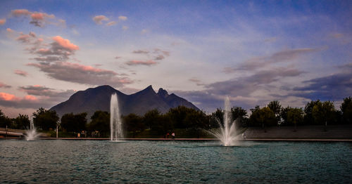 Fountain by lake against sky during sunset