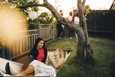 Smiling female friends relaxing on hammock at yard during sunset