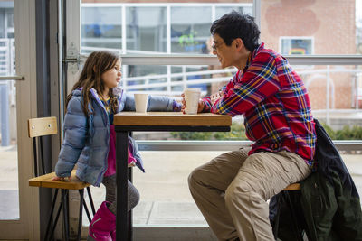 A little girl sits at a table by a large window with her happy father