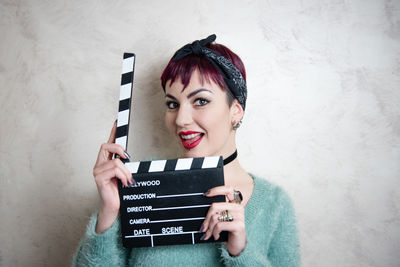 Portrait of smiling young woman holding film slate against wall