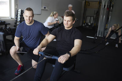 Smiling fitness instructor kneeling besides young man exercising on rowing machine at health club