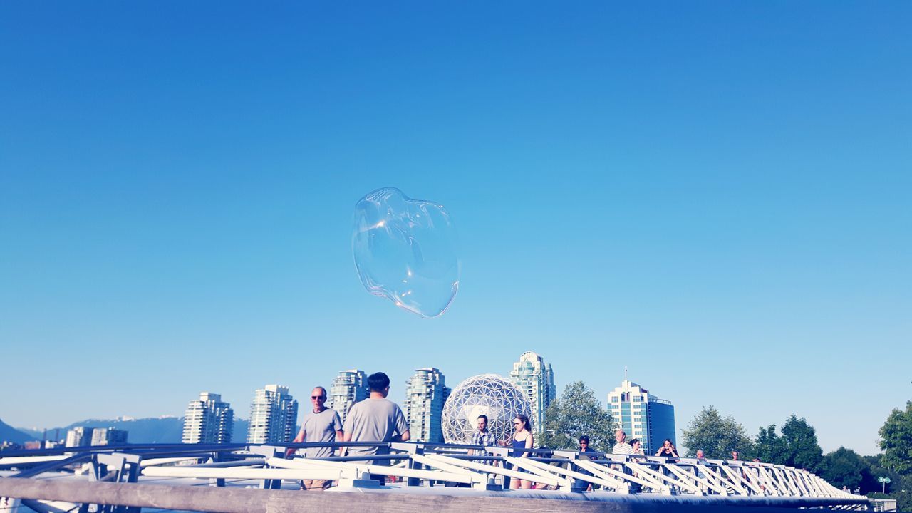 bubble, clear sky, blue, day, bubble wand, mid-air, outdoors, building exterior, built structure, architecture, real people, large group of people, fragility, nature, sky, close-up, people
