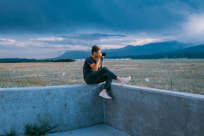 Young woman sitting on retaining wall against sky