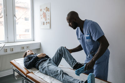 Male medical professional checking leg of teenage boy lying on bed in examination room at clinic