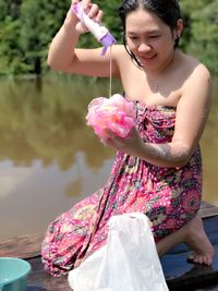 Smiling young woman pouring body lotion on loofah while kneeling at jetty over lake