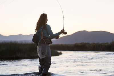 Side view of young woman fly-fishing while standing in owens river against mountains during sunset