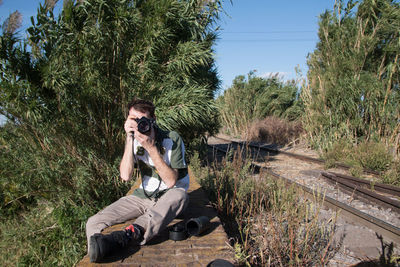 Man photographing while sitting by trees and track against sky