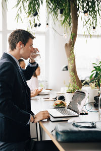 Businessman with bluetooth headphones drinking water while using laptop at office desk