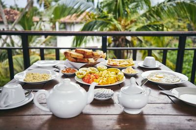 Breakfast ready to eat. eggs, tropical fruit, toasts and tea pot on wooden table against palm tree.