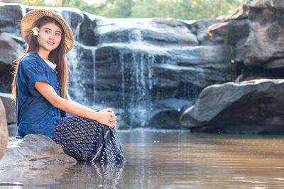 Young woman wearing hat while sitting on rock against waterfall