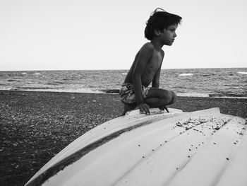 Boy sitting on upside down boat at beach against clear sky