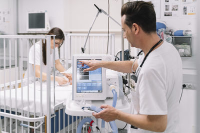 Side view of doctor male adjusting parameters medical device for artificial respiration in ward with nurse examining baby on background