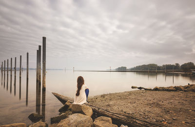 Rear view of woman relaxing on tree trunk by lake against cloudy sky