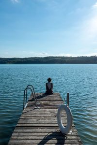 Rear view of man sitting on pier over lake against sky