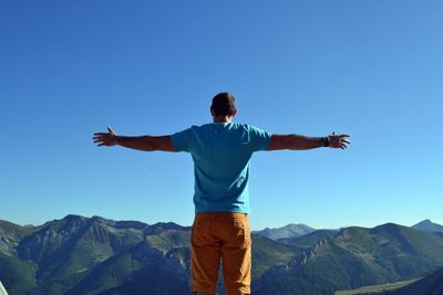 Rear view of man with arms outstretched standing by mountain against clear blue sky
