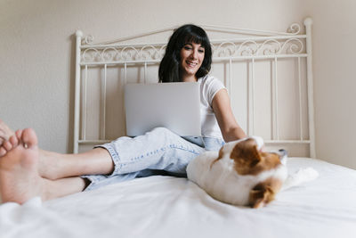 Young woman using laptop while puppy sleeping on bed