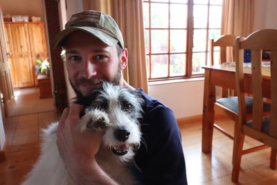 Portrait of young man carrying dog while standing at home