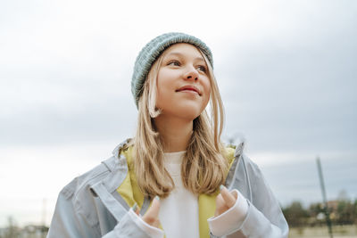 Thoughtful teenage girl wearing knit hat standing under sky