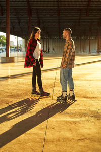 Full body of smiling man chatting to girlfriend during roller skating practice in urban park in sunny evening