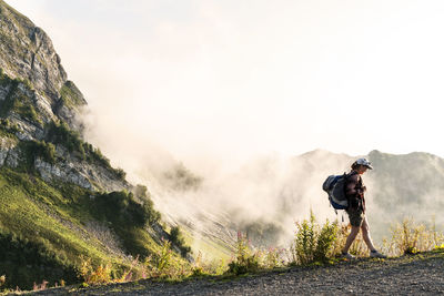 Young woman in cap and plaid shirt with backpack hiking in green mountains against clouds landscape