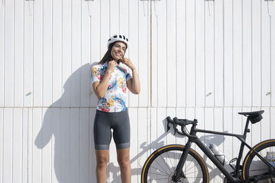 Happy cyclist adjusting helmet standing in front of white wall on sunny day