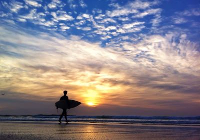 Silhouette of surfer walking on beach with surfboard during sunset