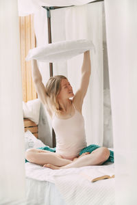 A beautiful smiling girl sits cross-legged on the bed and holds a pillow on her outstretched arms. 