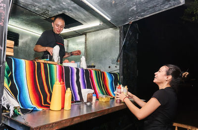 Positive ethnic woman with sauce bottles standing against food truck and looking at content male colleague at counter at night