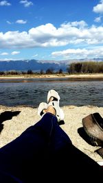 Low section of man sitting by lake against sky