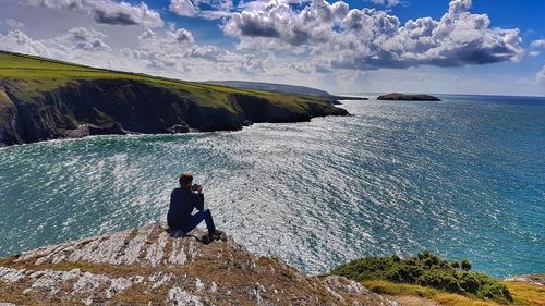 Man sitting on cliff by sea against sky