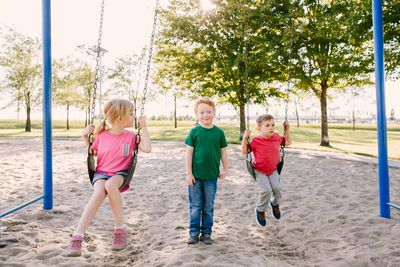 Happy smiling little girl and boys friends swinging on swings at playground outside on summer day. 