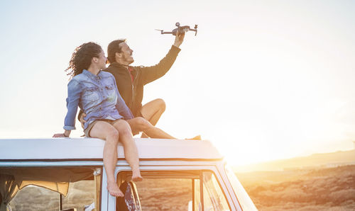 Man with woman holding drone while sitting on van against sky