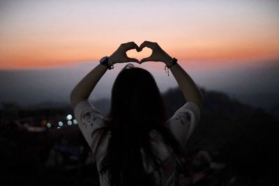 Rear view of woman making heart shape over head against sky during sunset