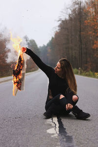 A woman sitting on the empty road, holding a burning paper in hand 