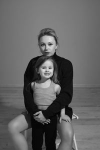 Mother in a black sweater hugs her five-year-old daughter sitting on a chair in the room in front