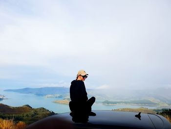 Rear view of young woman sitting on car roof against sky