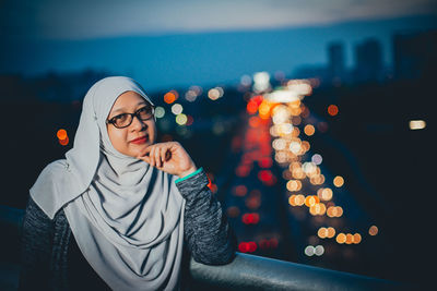 Portrait of woman wearing hijab by railing against illuminated city