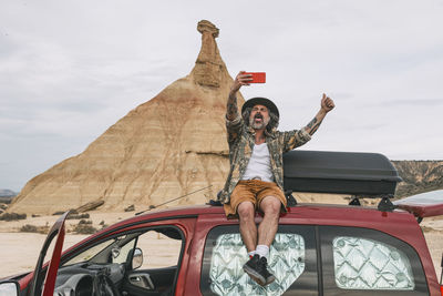 Tourist sitting on top of a van using a mobile camera to take one with the rocky landscape. person