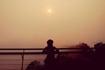 Rear view of silhouette woman standing by railing against sky during sunset